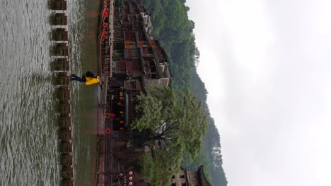 tracking shot of a man in yellow raincoat crosses tuojiang on stepping stones in fenghuang on a cloudy day, china