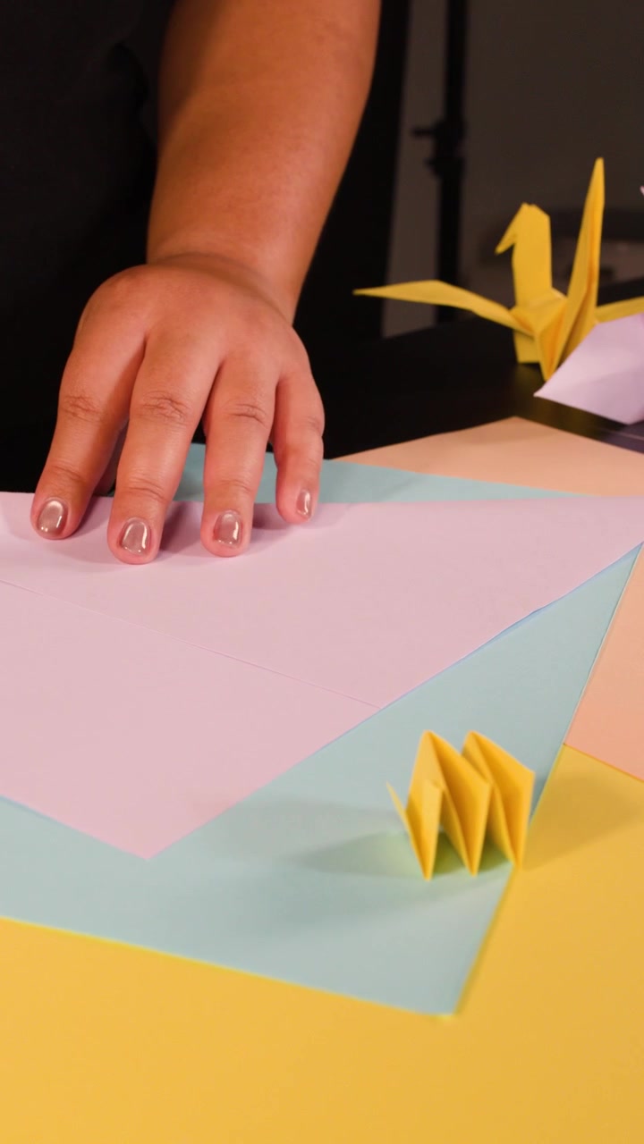 Una persona piega carta rosa su una scrivania colorata con gru e forme di origami finite.