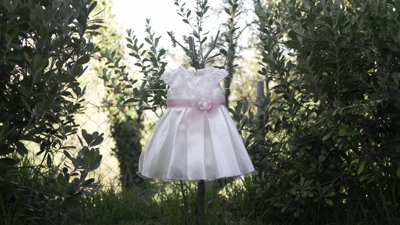 Baby dress with floral belt hanging between green bushes outdoors, soft light