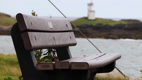 a wooden bench overlooking a distant lighthouse across the sea, surrounded by grassy terrain.