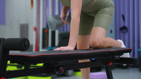 vista parcial de una mujer realizando un ejercicio de fila de pesas, descansando la mano y la rodilla en el banco de entrenamiento para apoyo, centrándose en la activación muscular, la estabilidad y la rutina de entrenamiento de movimiento controlado