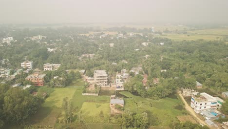 drone lifts up and moves backward, unveiling a serene rural village in bangladesh with scattered houses and lush greenery.
