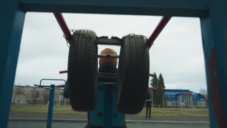 back view of a coach doing pull-ups on workout equipment while two boys stand in the background, with someone in pink riding an electric bike on a track nearby