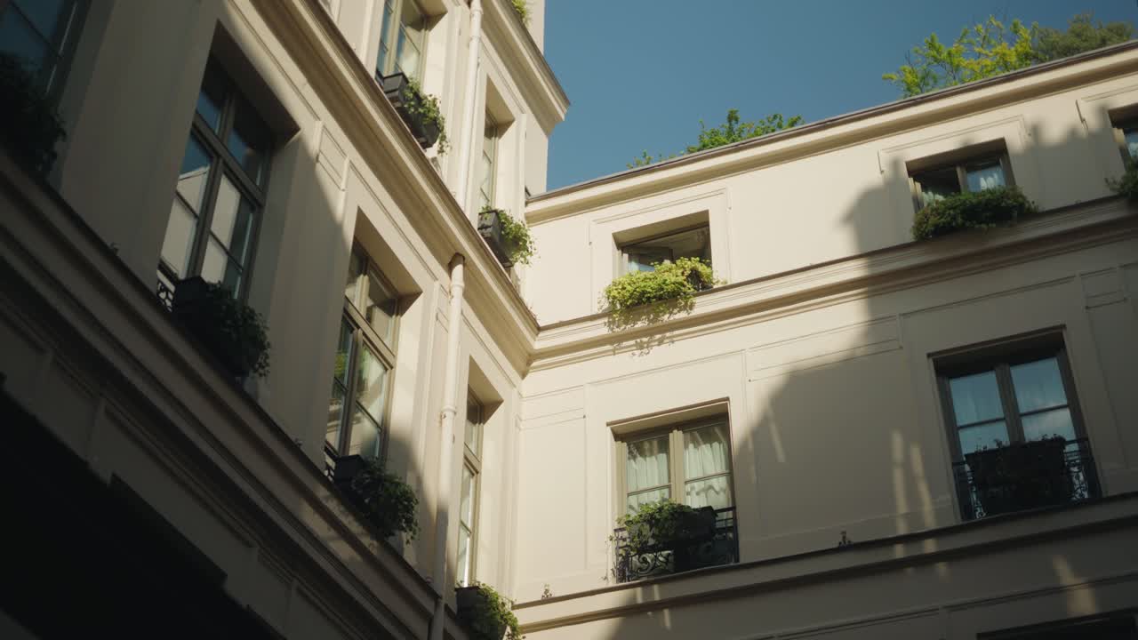 Inclinação ascendente lenta de uma fachada de pátio interno ensolarado com janelas altas, varandas e folhagens em vasos em um elegante cenário arquitetônico urbano.