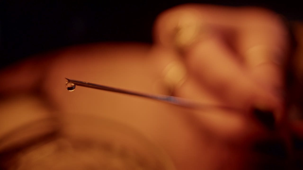 A close-up view of a liquid drop on a needle held by a hand wearing gold rings.