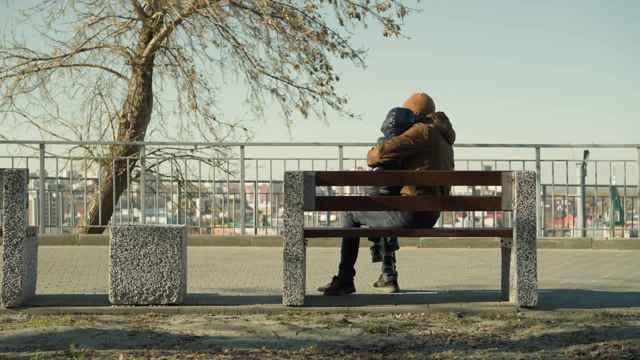 A father gently carries his son on his leg while they sit together on a bench, the father holds him affectionately as he seat sideways on the bench with a cityscape in view