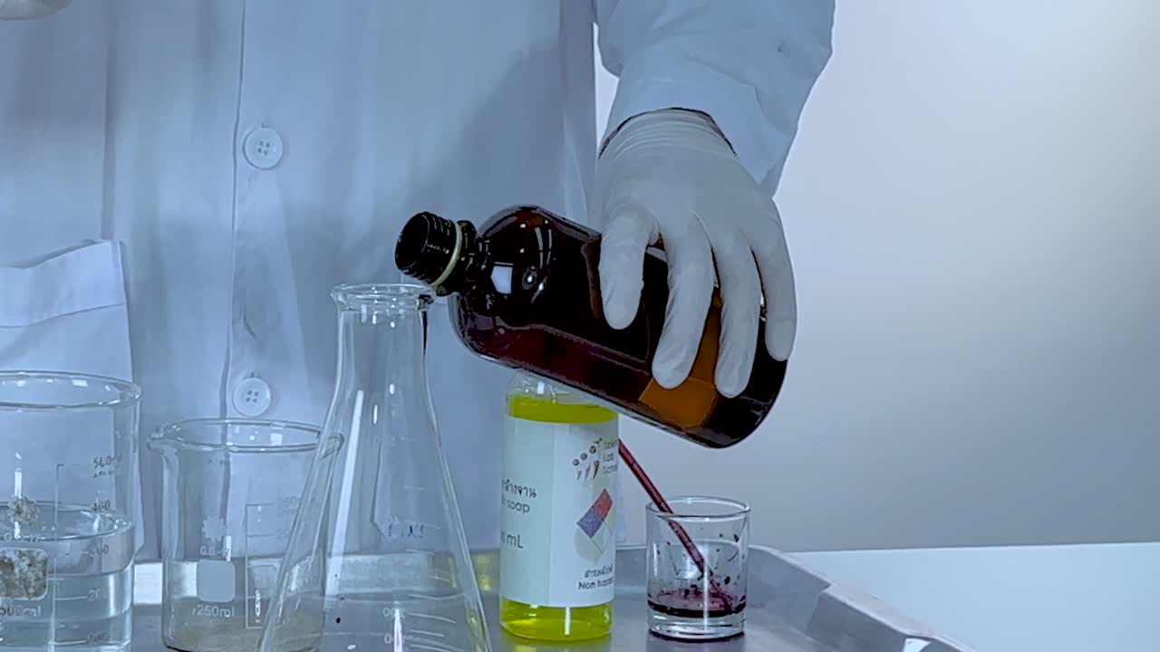 A scientist pours liquid from a brown bottle into a flask in a lab setup.