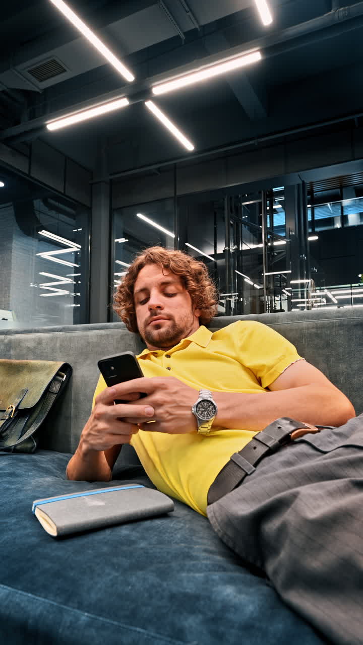 Man in yellow shirt lying on a couch and scrolling on his phone. Vertical