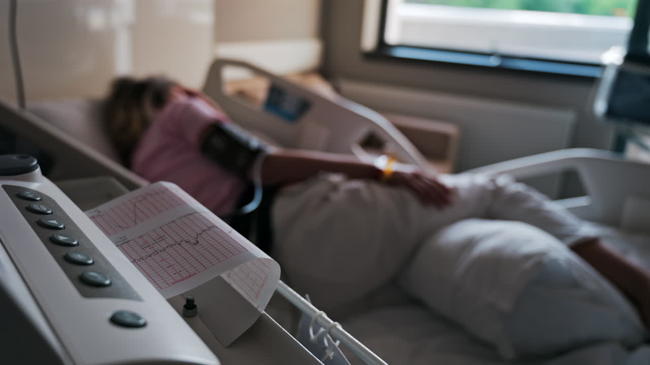 Close-up of a fetal monitor in a doctor's office, displaying fetal heart rate and uterine contraction data during a prenatal check-up with a blurred view of a pregnant woman lying in a hospital bed