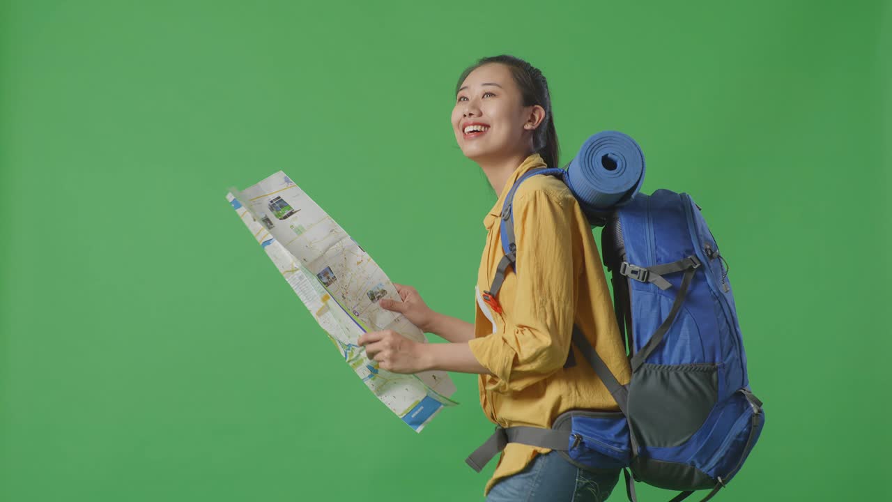 vista lateral de una excursionista asiática con mochila de montañismo mirando el mapa y luego mirando a su alrededor mientras camina en el fondo de la pantalla verde en el estudio