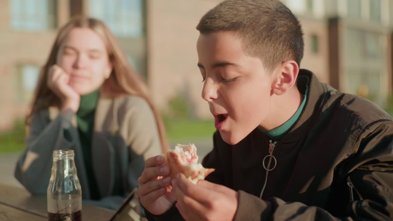 Boy enjoying bite of pizza outdoors with relaxed smile while woman sits beside him with closed eyes under warm sunlight, juice bottle and sandwich pack resting on wooden table in casual urban setting
