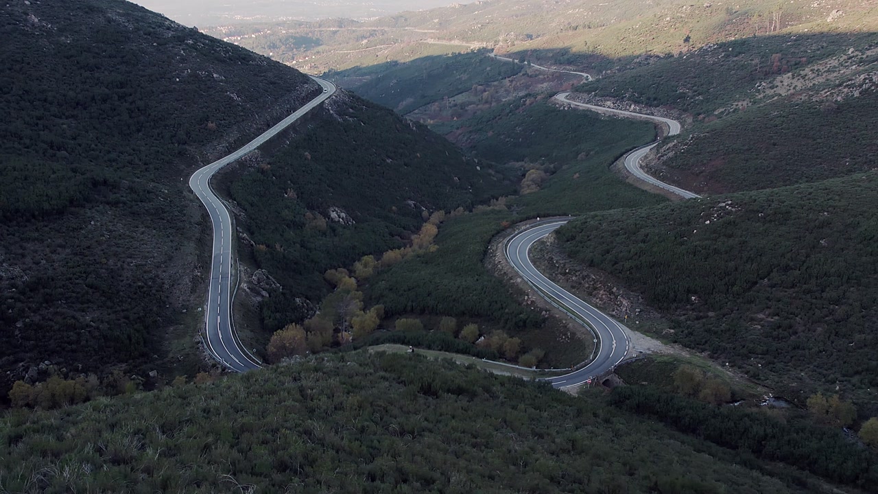 N338 mountain road with multiple switchbacks winding through steep valley in Serra da Estrela, Central Portugal, showing shrub vegetation, riparian trees and rugged high-altitude terrain, drone orbit