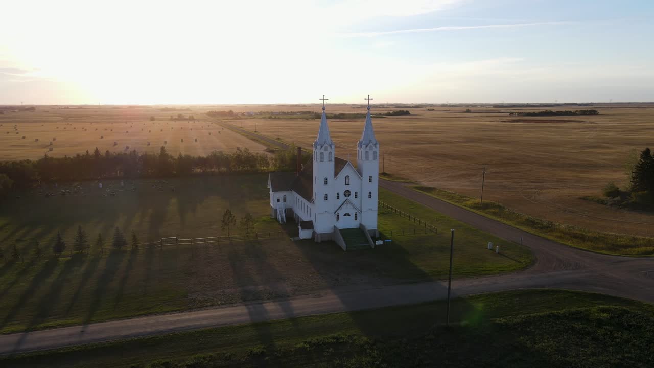 Aerial circle shot of roman catholic church in north American prairie during sunset. White building with blue roof on steeples.