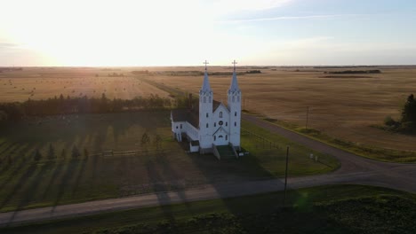aerial circle shot of roman catholic church in north american prairie during sunset. white building with blue roof on steeples.