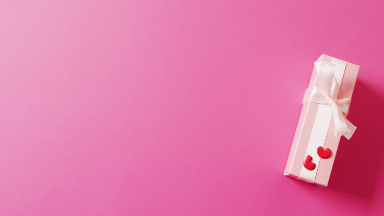 Video of pale pink gift box with white bow and red hearts, on pink background with copy space