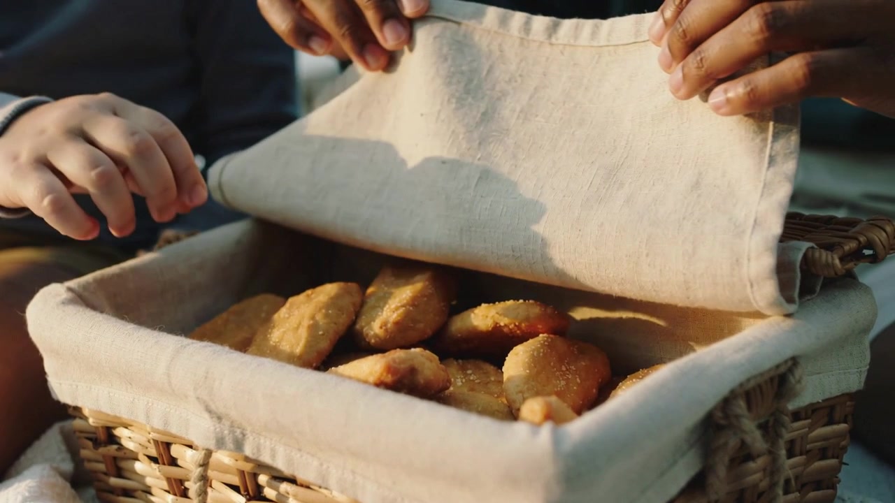 Lifting cloth, adult hands revealing wicker basket at picnic, offering snack as child grabbing one
