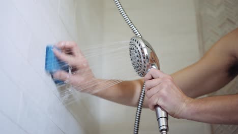 Weißer-Mann-Schrubbt-Die-Nähte-Seiner-Badezimmerwand-Mit-Einem-Blauen-Schwamm-Und-Einem-Verchromten-Duschkopf