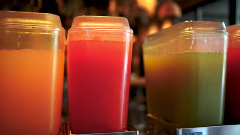 Orange, red and green juices are displayed in transparent plastic containers on a metal support, ready to be served to restaurant customers