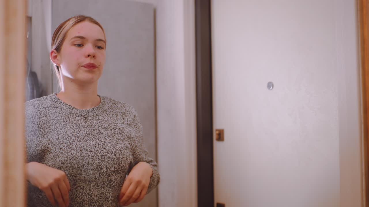 Young woman in gray sweater adjusting hair while checking reflection in hallway mirror before leaving home, showing grooming, style awareness, and thoughtful preparation