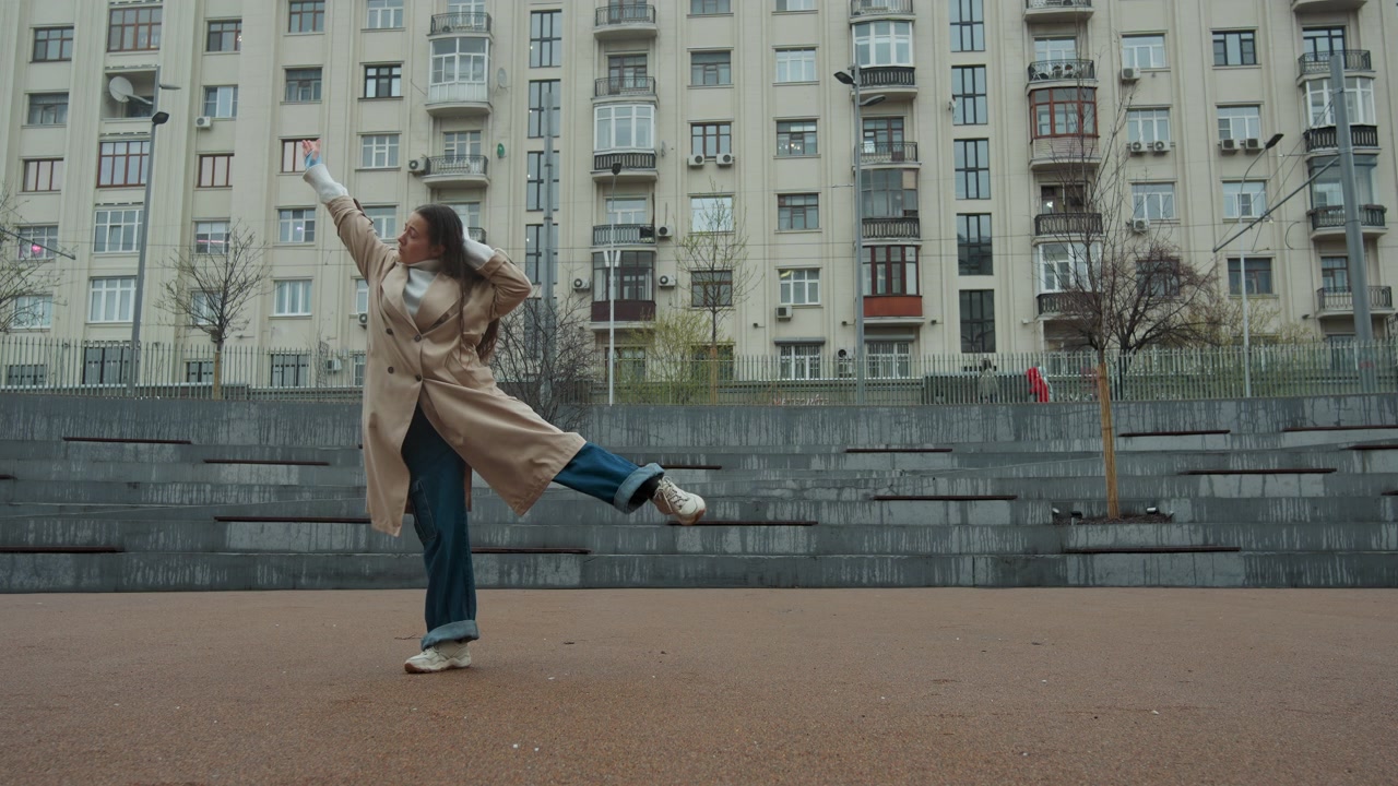 Young woman performs dance routine in urban outdoor setting wearing beige trench coat and blue jeans. Woman moves gracefully across frame with fluid arm gestures against backdrop of tall