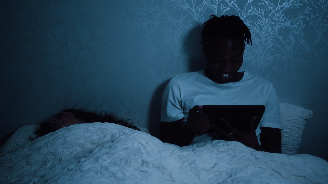 hombre sentado en la cama tarde en la noche sonriendo mientras mira el dispositivo tableta y su pareja duerme a su lado