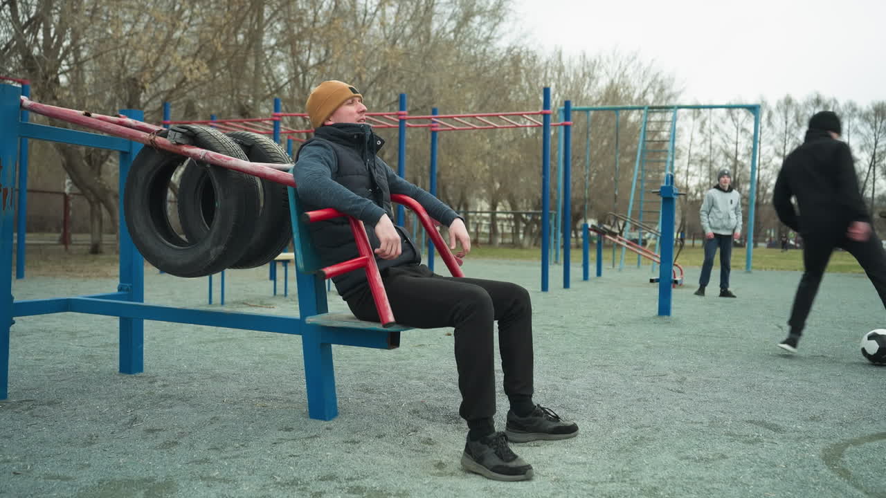 A coach leans back on workout equipment, casually shaking his leg, in the background, two boys are actively playing football