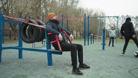 a coach leans back on workout equipment, casually shaking his leg, in the background, two boys are actively playing football