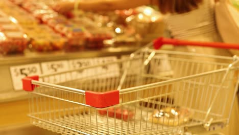 a woman selects groceries in a well-lit supermarket aisle, pushing a shopping cart filled with items