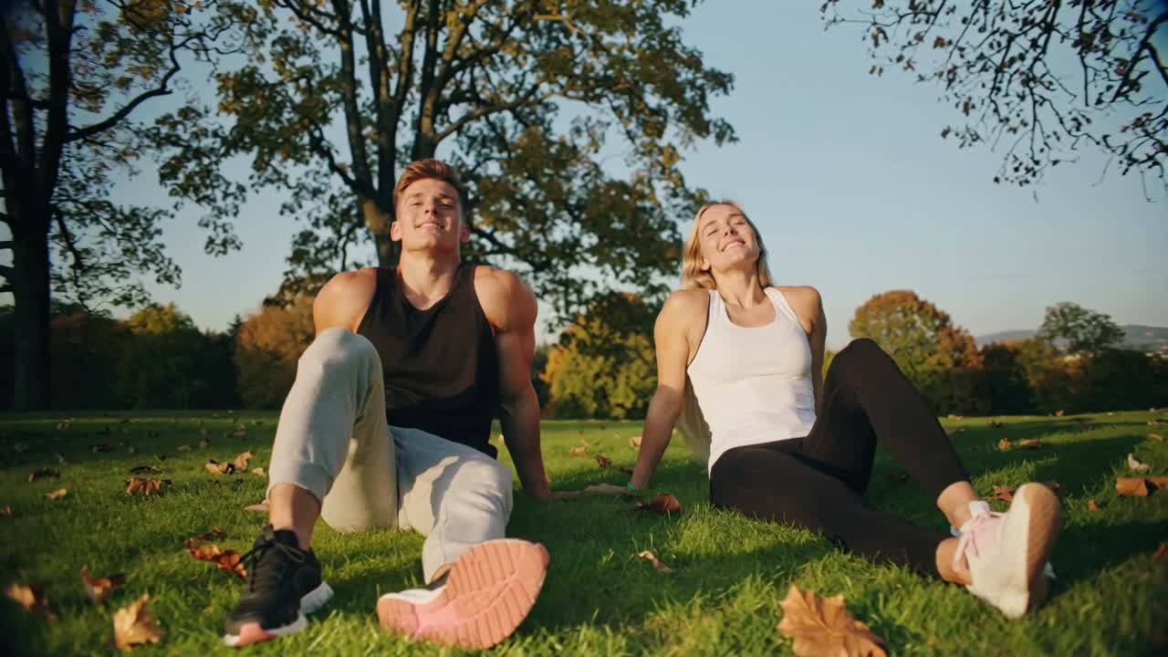 Casal sentado descansando em uma encosta gramada do parque após a corrida, alongando as pernas, usando regatas e tênis.