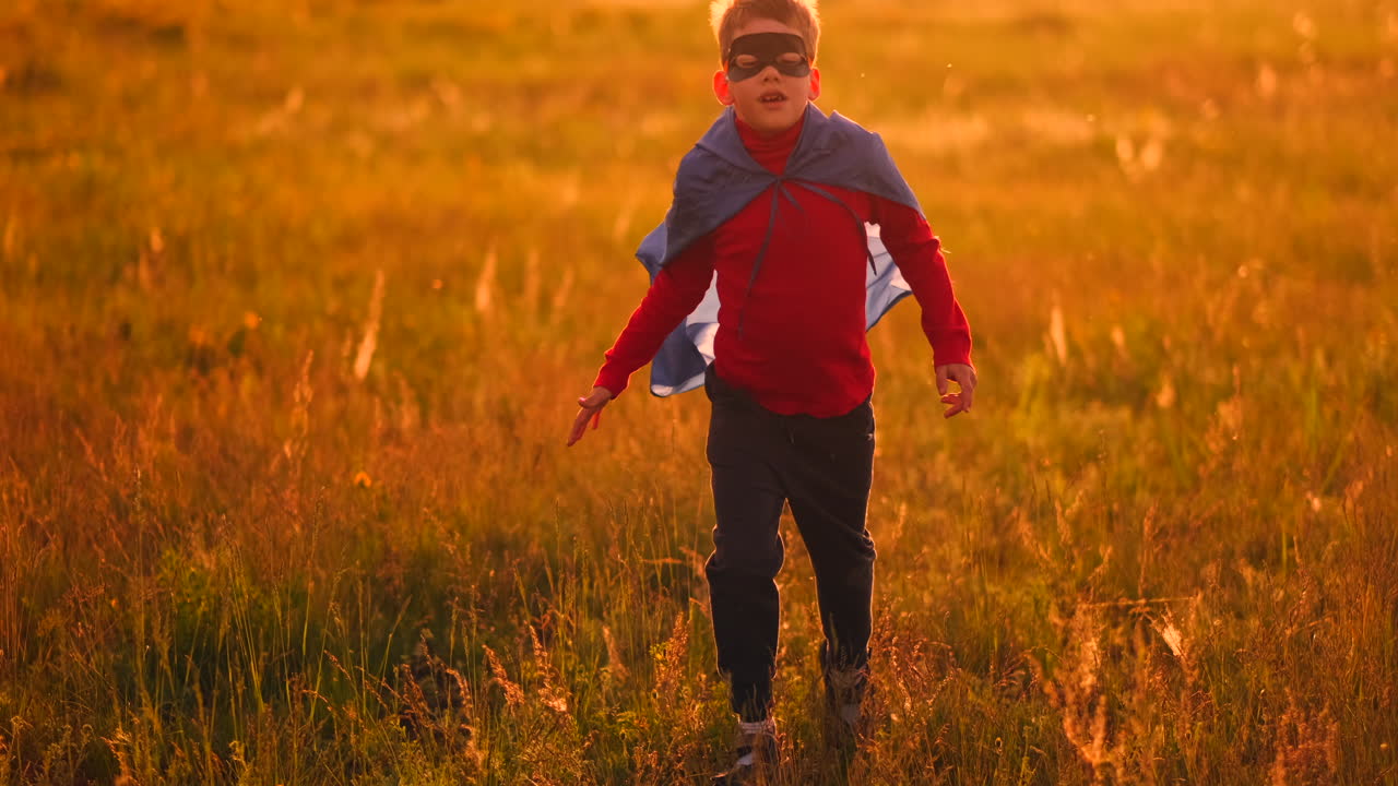 un niño con un traje y una máscara de superhéroe con una capa roja corre a través del campo al atardecer en la hierba soñando e imaginándose a sí mismo un héroe.