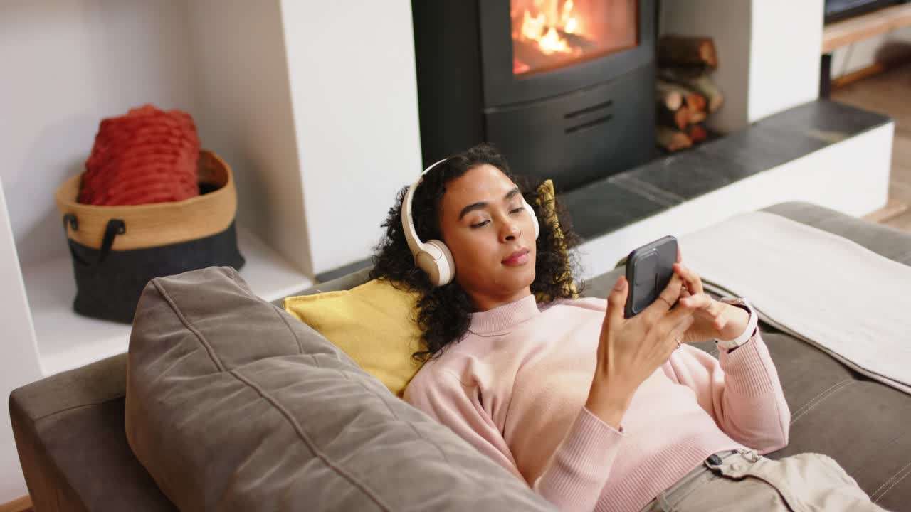 Persona non binaria che prende lo smartphone e scorre i contenuti su un divano grigio vicino a una stufa accesa