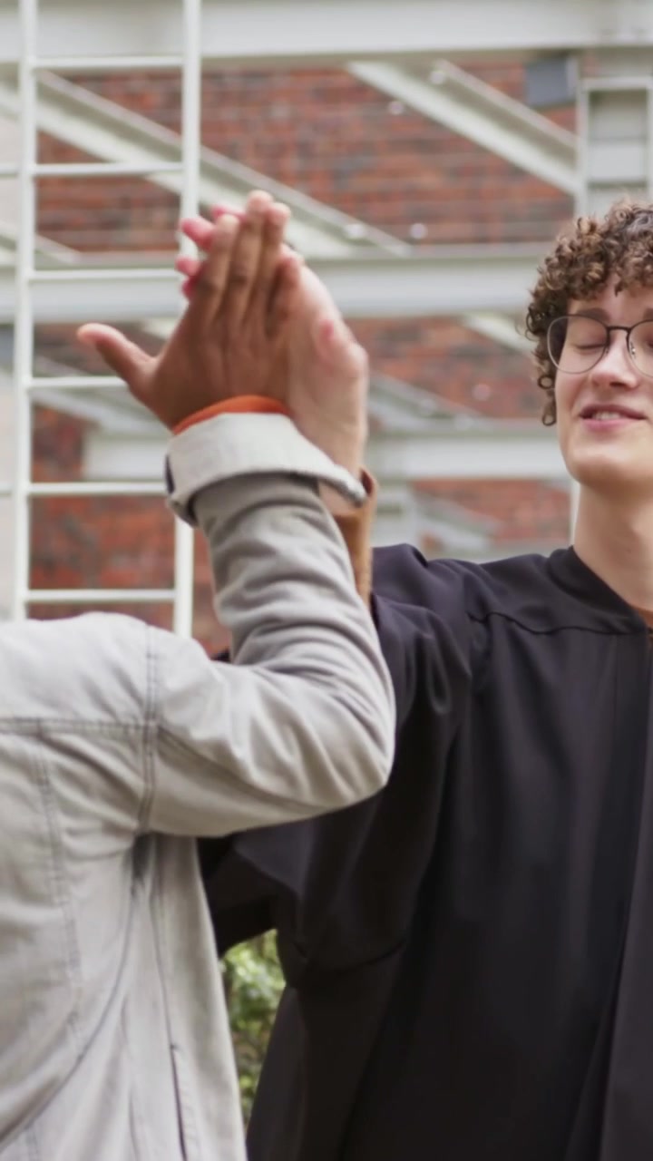 Video verticale: Laureato sorridente in toga con nappa gialla, che dà il cinque a un amico dopo la cerimonia della nappa nel campus.