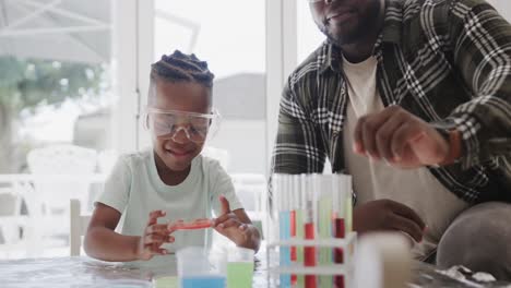 african american father and son sitting at table holding test tubes with liquid, in slow motion