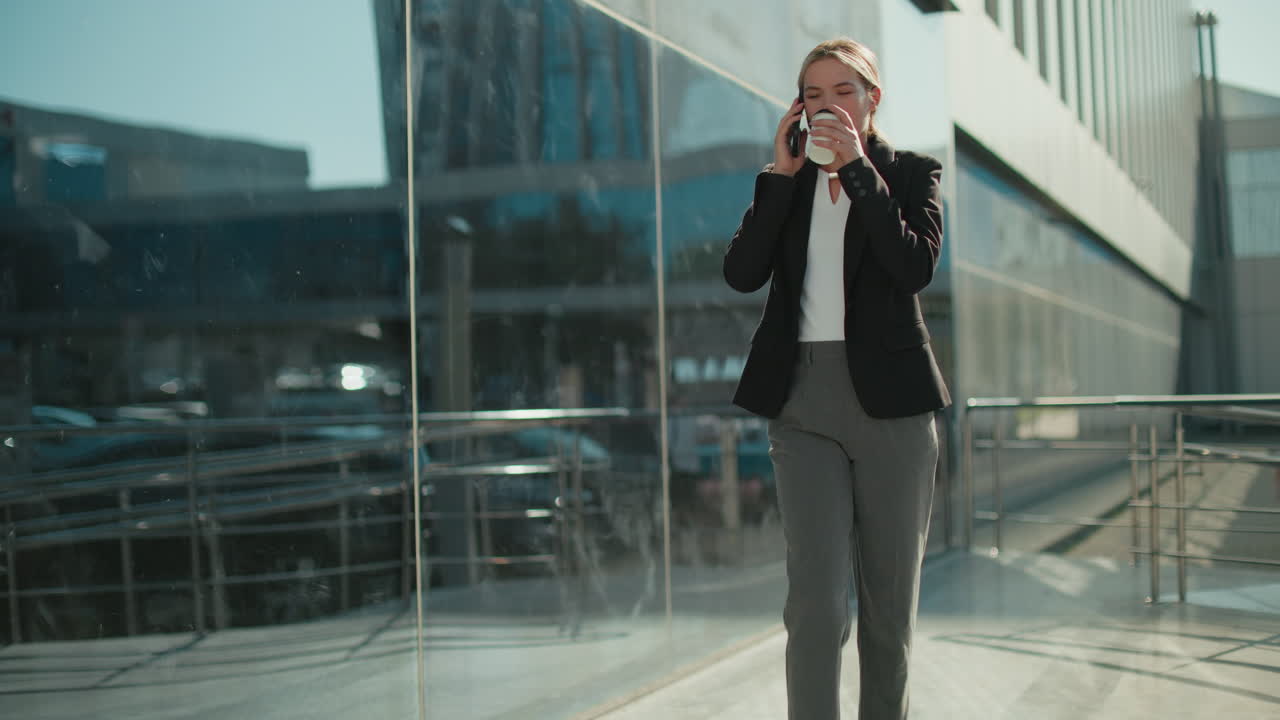 Professional lady walking confidently with coffee in hand on phone call beside iron railing, reflected on glass building exterior with people in background