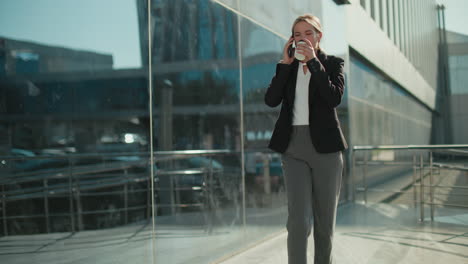 Professional lady walking confidently with coffee in hand on phone call beside iron railing, reflected on glass building exterior with people in background