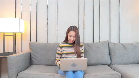 a young woman sitting on a couch works on her laptop suddenly reacts to her screen by raising her arms in success