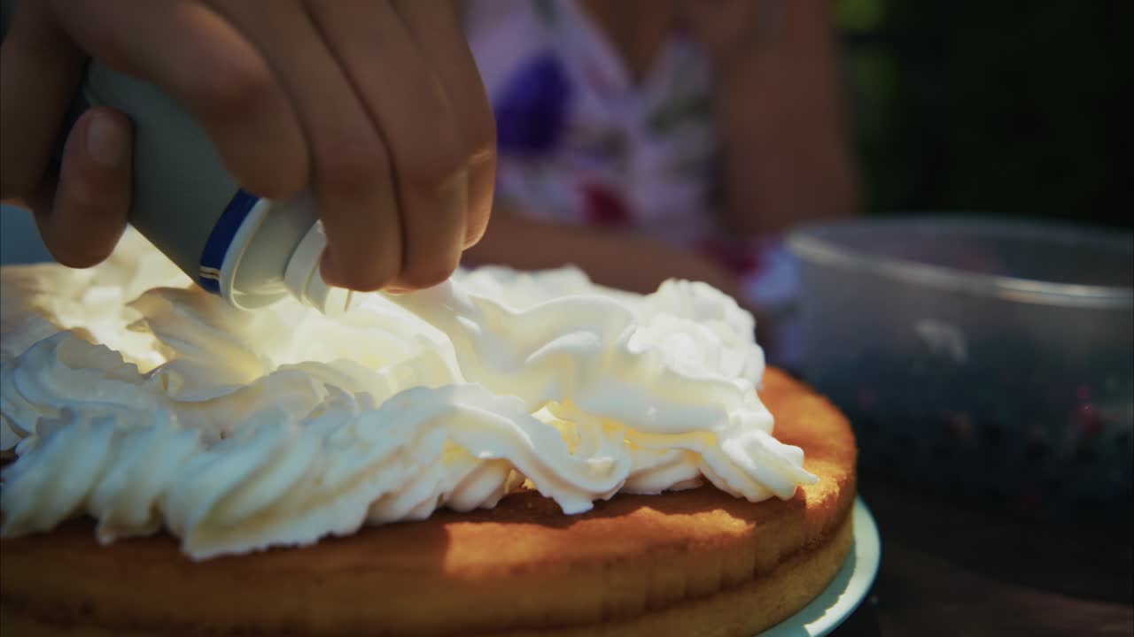 Hand applying whipped cream on a freshly baked cake in a sunny outdoor setting with vibrant colors