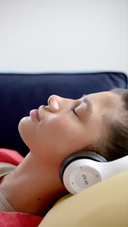 vertical video of smiling biracial teenage girl wearing headphones lying on couch, slow motion
