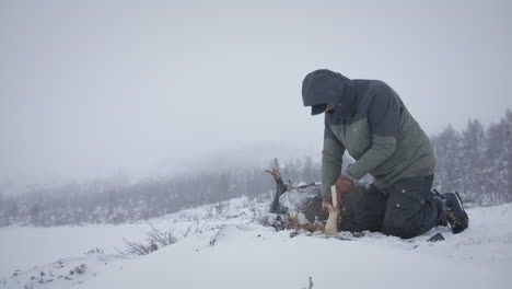 a person in winter clothing kneels in snow while chopping wood, surrounded by a foggy, frozen landscape.