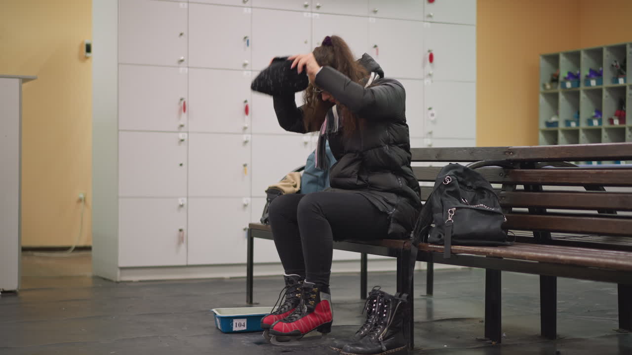 Girl wearing glasses, black jacket, striped scarf, and red ice skates sits on bench in locker room tying hair, black boots and backpack nearby, preparing for skating with focus and natural candid expression
