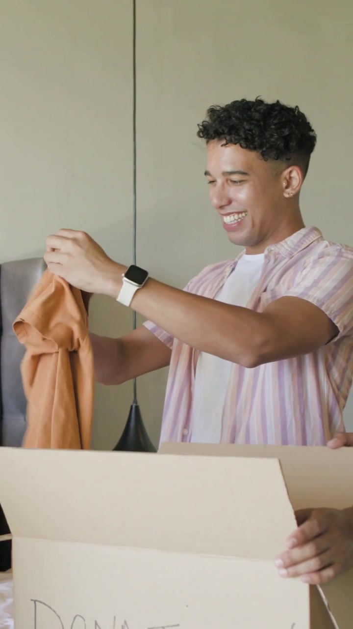 Vertical video: Pulling orange shirt, man in pink shirt folding and packing DONATIONS boxes on bed