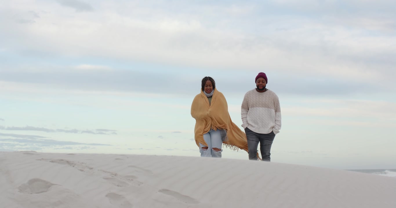 African American couple cresting dune and walking down toward camera, woman holding yellow blanket
