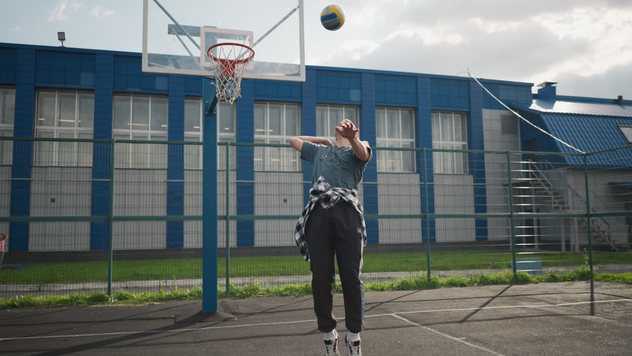el deportista lanza la pelota de voleibol, salta y la golpea mientras está de pie en la cancha de baloncesto, el edificio azul y el fondo cercado hacen un tiro dinámico y enérgico lleno de acción y movimiento deportivo
