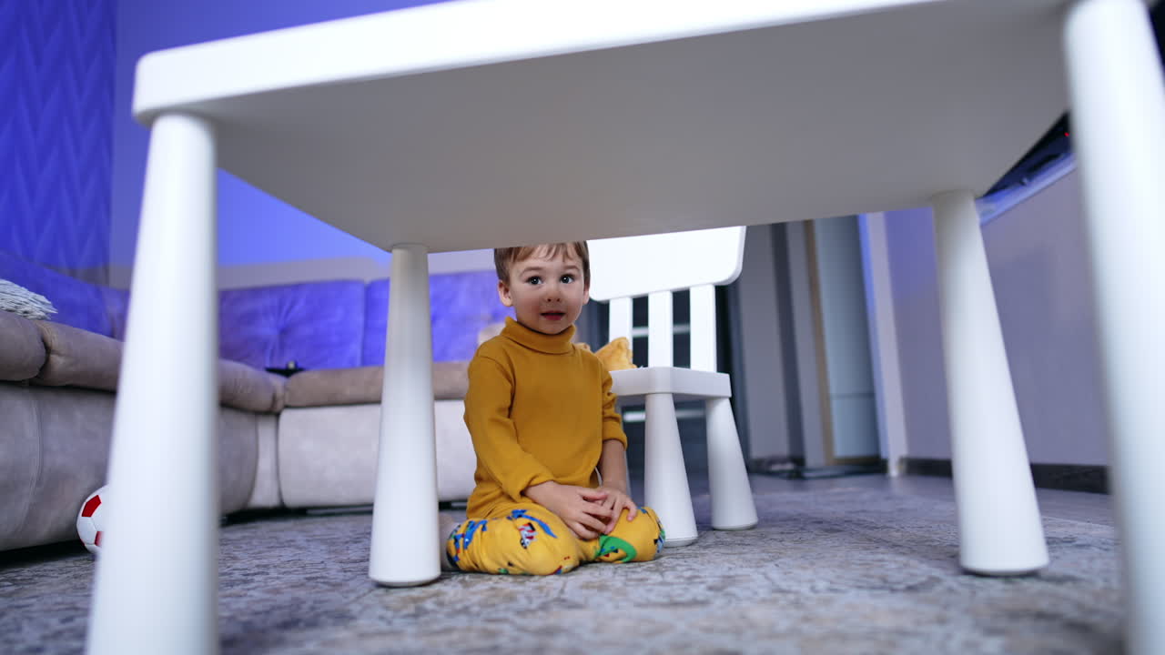 Cute two year old toddler sitting on his knees on the floor near the desk. Baby boy in yellow clothes crawls by the floor.