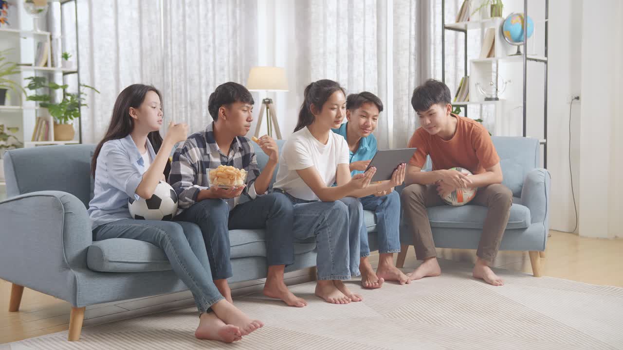 아시아 십대들이 집에서 태블릿으로 축구 경기를 즐기고, 먹고, 보고 있습니다.