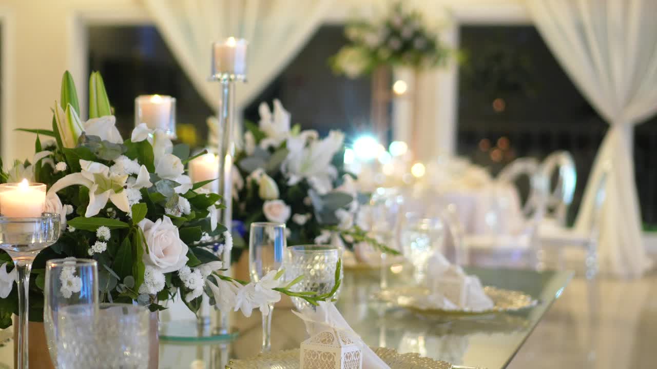 Tavolo di vetro decorato con centrotavola di fogliame naturale e rose bianche, candelabri e cristalleria per ricevimento di matrimonio