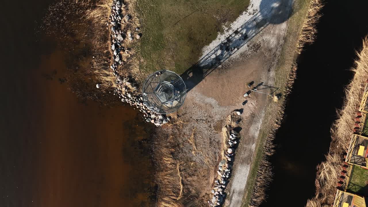 An observation tower stands next to a body of water, with a couple walking on top. Dreverna village, Lithuania.