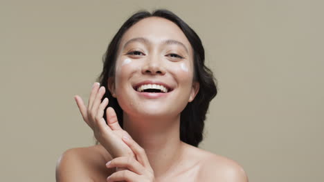어두운 머리카락을 가진 행복한 아시아 여성이 베이지색 배경에 얼굴에 크림을 바르며, 느린 움직임