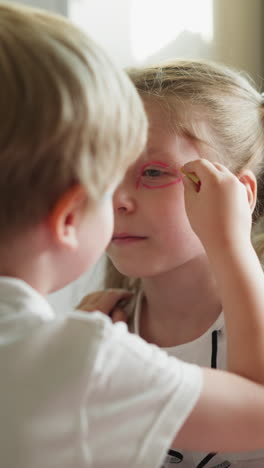 younger brother paints sister face with red pencil slow motion. siblings have fun playing together at home. little artist draws glasses closeup