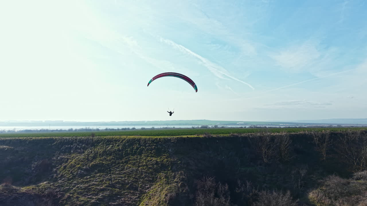 Flying over green hills near the river while enjoying paragliding during bright daylight with clear skies